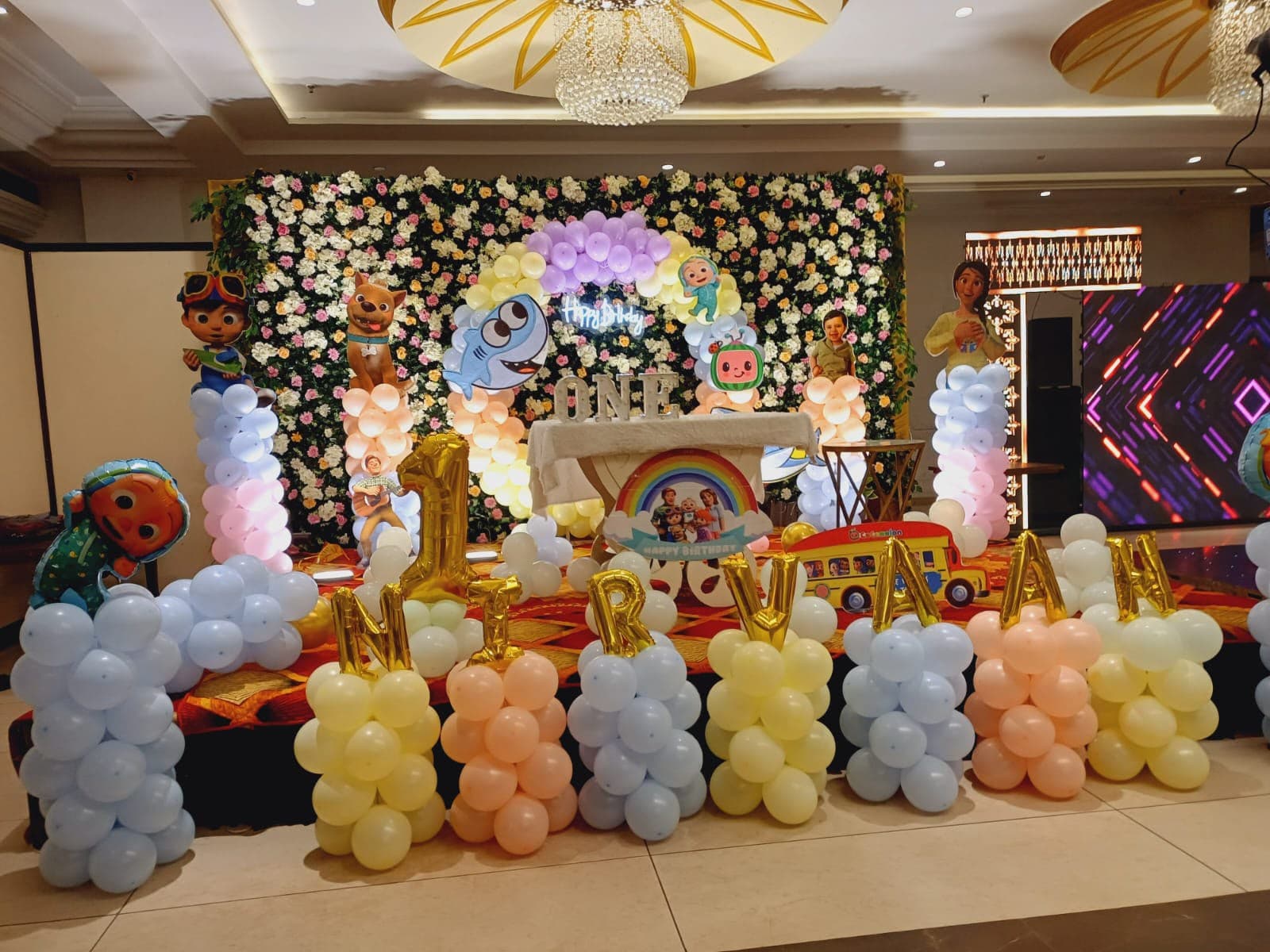 First birthday stage with a white floral wall, pastel balloon columns and cartoon cut-outs